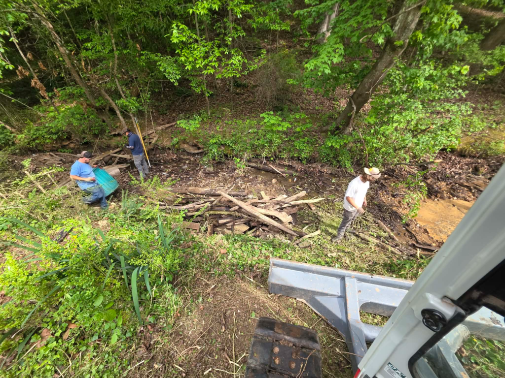 Three crew members clearing accumulated tree debris and wooden boards from creek bed – White's Tree Service Knoxville TN
