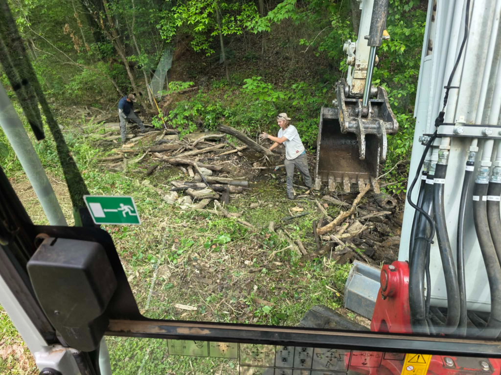 Excavator removing large logs from creek bed while crew member guides the bucket – White's Tree Service Knoxville TN